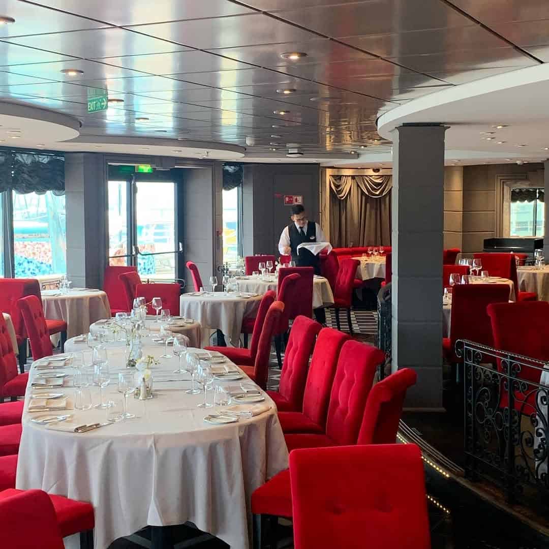 Elegant restaurant on MSC Fantasia Dining, featuring red chairs, round tables set for dining, and a staff member preparing in the background.