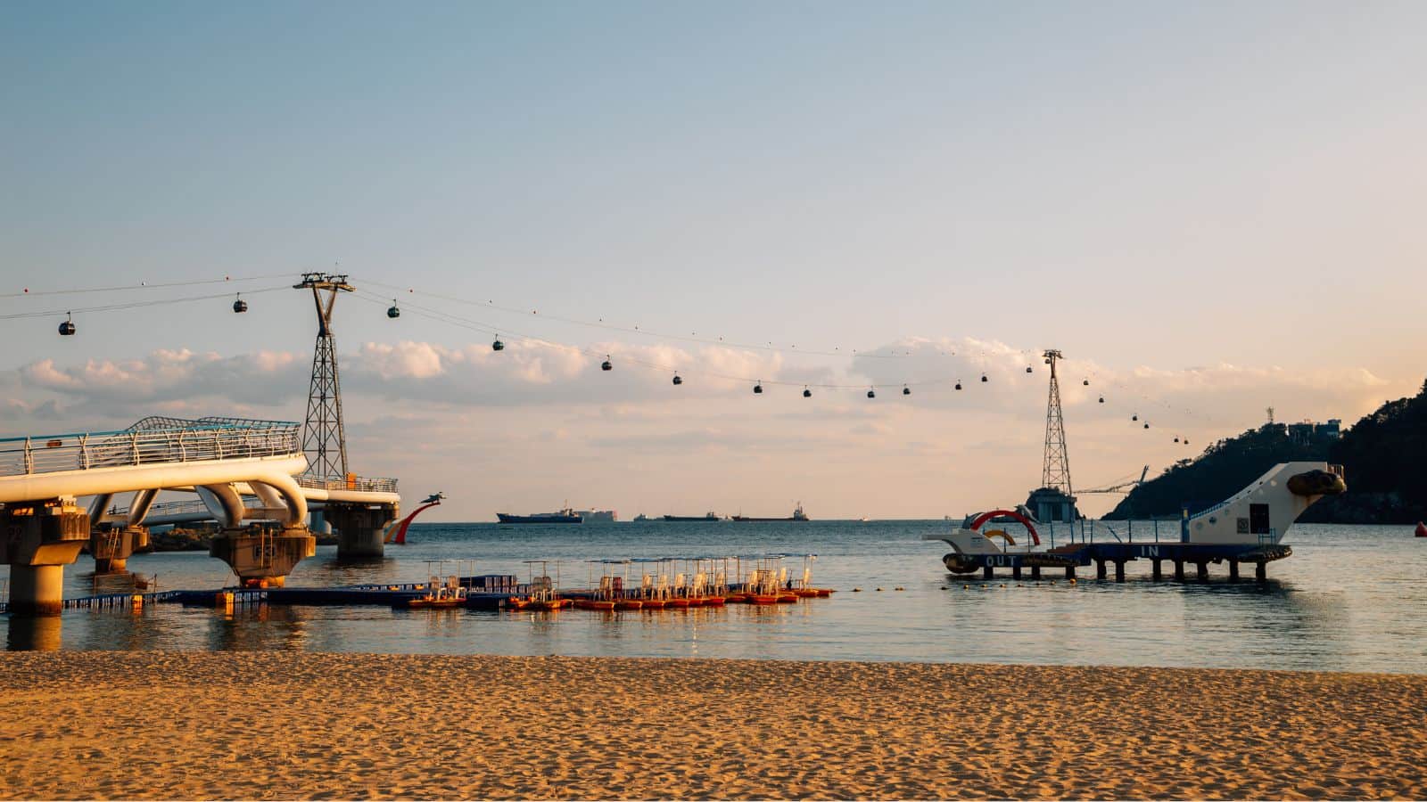 A sandy beach with piers, floating docks, and cable cars crossing above the calm sea at sunset.