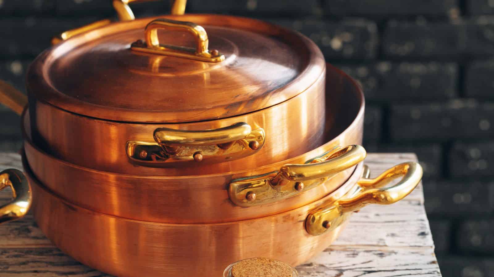 Stacked copper pots with gold handles sit on a rustic wooden surface against a dark brick background.