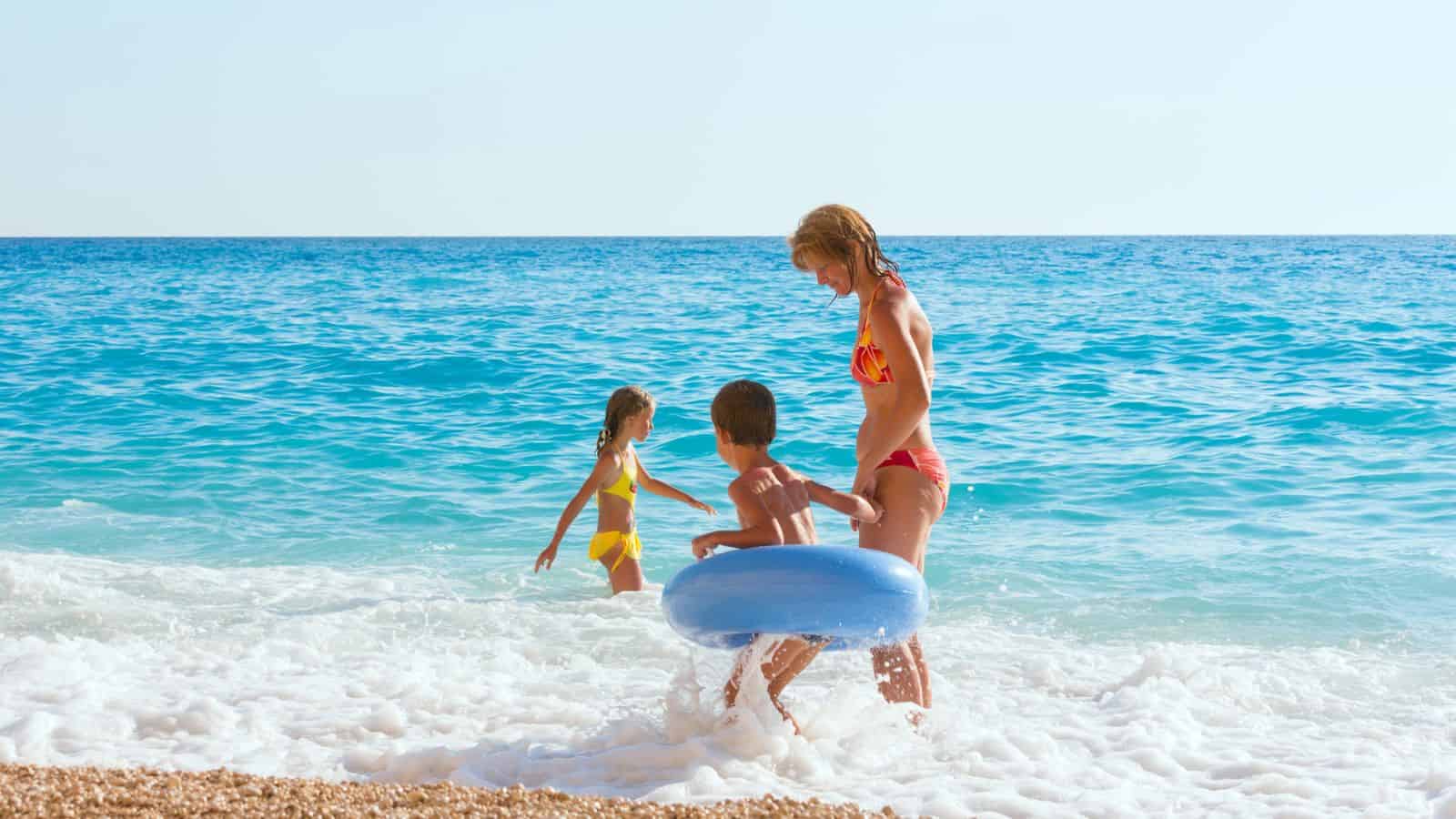 An adult and two children play at the edge of the sea on a sandy beach; the adult holds a child in a blue float ring while another child stands nearby in the shallow water. The sky is clear and sunny.