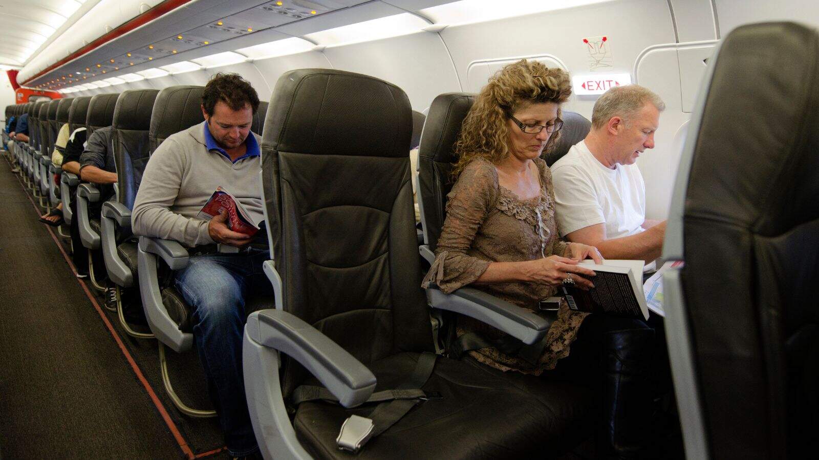 Several people sit in airplane seats, each reading a book or magazine. The cabin is well-lit, and the passengers appear focused on their reading material. The airplane aisle and emergency exit sign are visible.