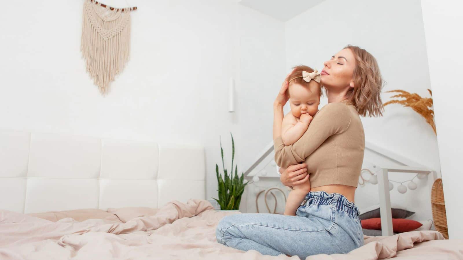 Woman sitting on a bed, hugging a baby with a bow headband in a cozy, softly lit bedroom.