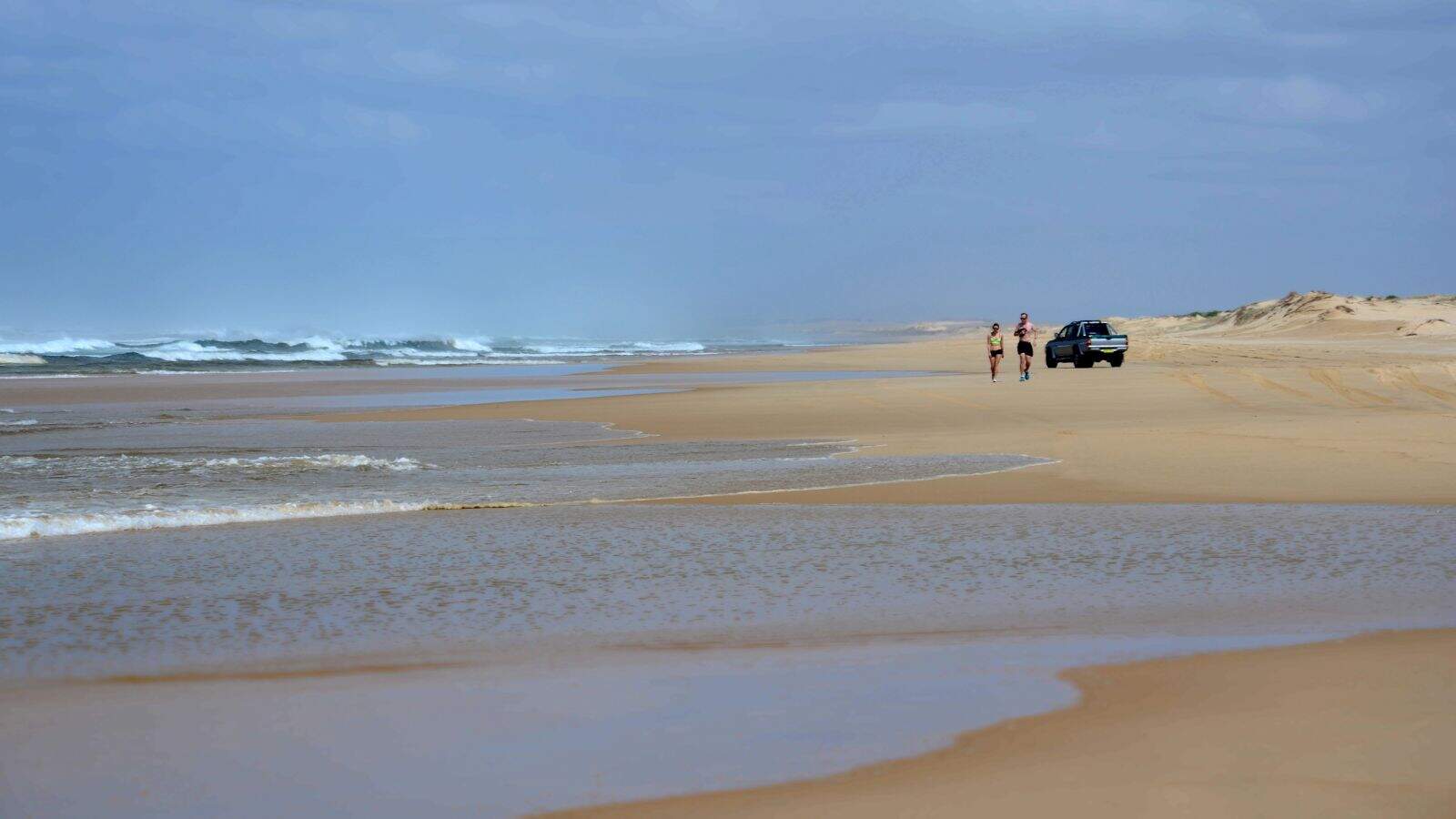 A sandy beach with two people walking near the shoreline and a parked vehicle farther behind them. Waves are visible on the left, and sand dunes extend into the distance on the right under a cloudy sky.