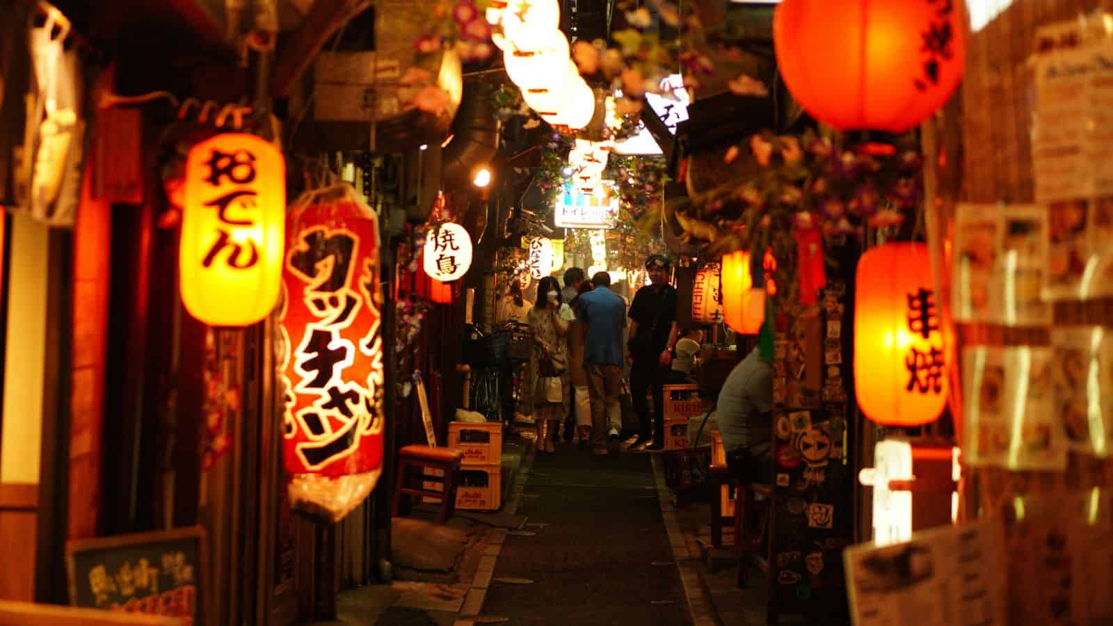 Narrow alley in Japan lit by red lanterns, with people dining and walking among cozy restaurants at night.