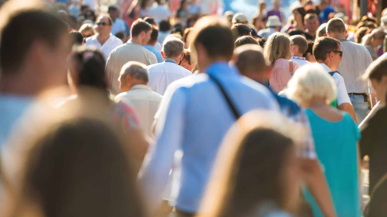 A large crowd of people walking outdoors in sunlight, seen from behind, with many out of focus.