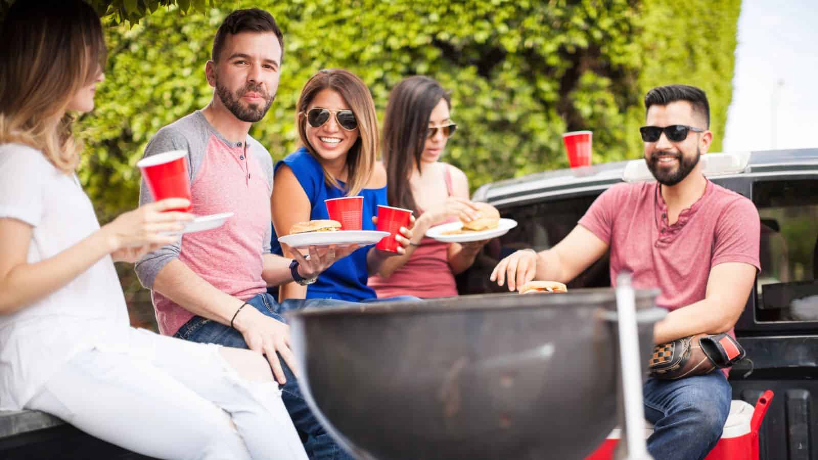 Five people sit and stand around a grill and pickup truck, holding red plastic cups and plates with food, outdoors on a sunny day near green hedges. They are smiling and appear to be at a casual gathering.