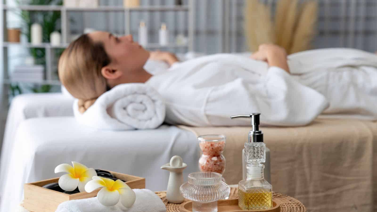 Woman in a white robe lying on a spa bed with oils and spa items in the foreground.