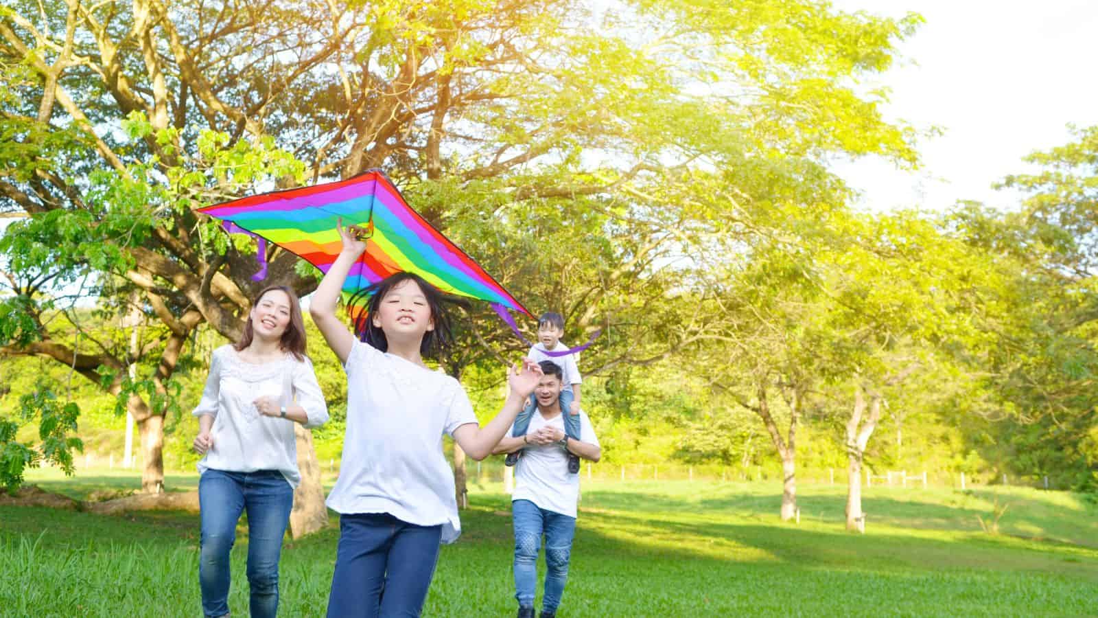 A family of four is outdoors on a grassy field. A child in front holds a colorful kite, followed by a woman, a man carrying a younger child on his shoulders, and trees and sunlight in the background.