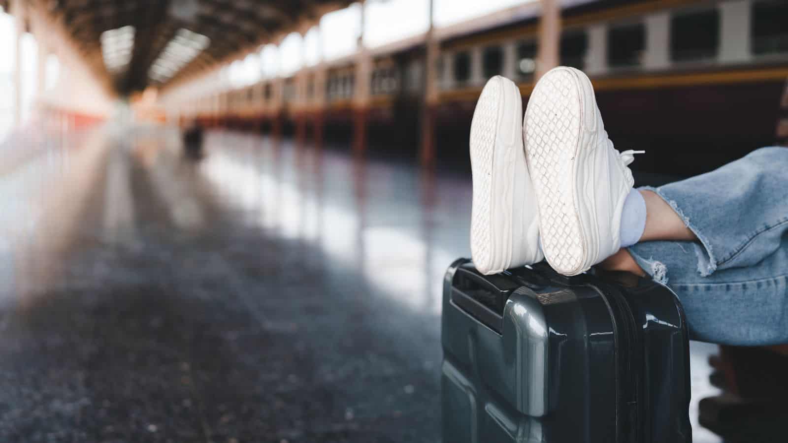 A person wearing white sneakers and blue jeans rests their feet on a black suitcase at a train station platform, embracing the excitement of train travel with blurred train carriages in the background.