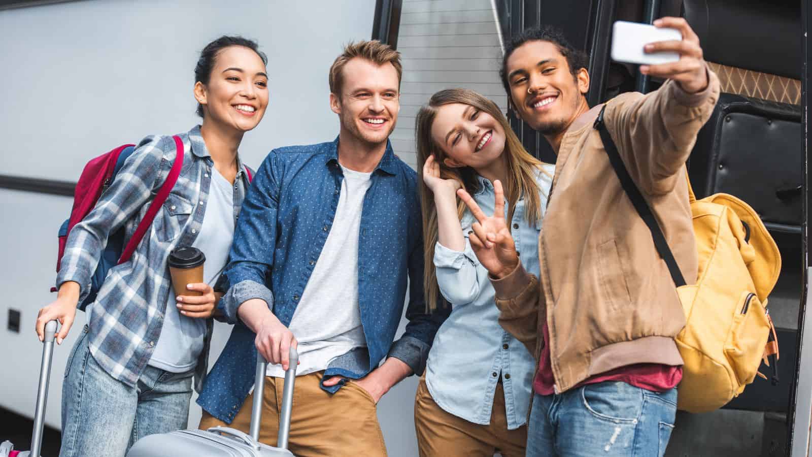 Four young adults stand in front of a bus, smiling and posing for a selfie. Two hold suitcases, one holds a coffee cup, and all appear to be ready for travel with backpacks and casual clothing.