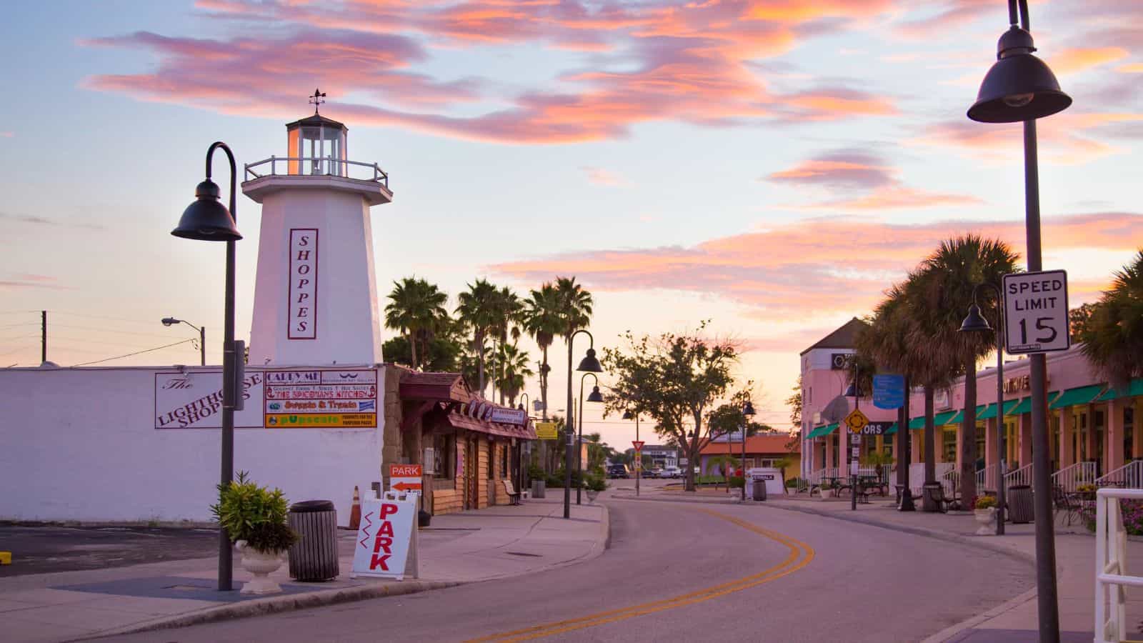 A quiet street in one of those charming American towns curves past a white lighthouse-style building labeled “Shoppes,” with palm trees, shops, street lamps, and a 15 mph speed limit sign under a colorful sunset sky.