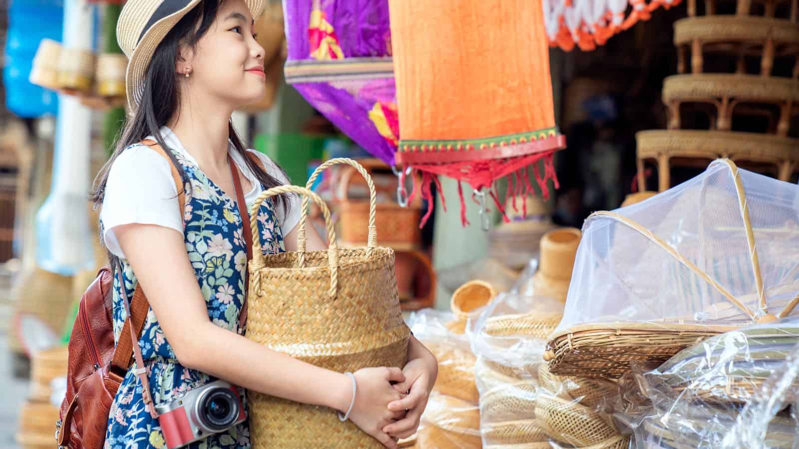 Young woman in a hat shopping at an outdoor market, holding a woven basket and smiling.