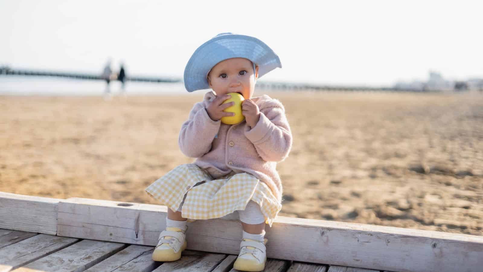Baby in a blue hat and pastel clothes sits on a boardwalk at the beach, eating a yellow apple.