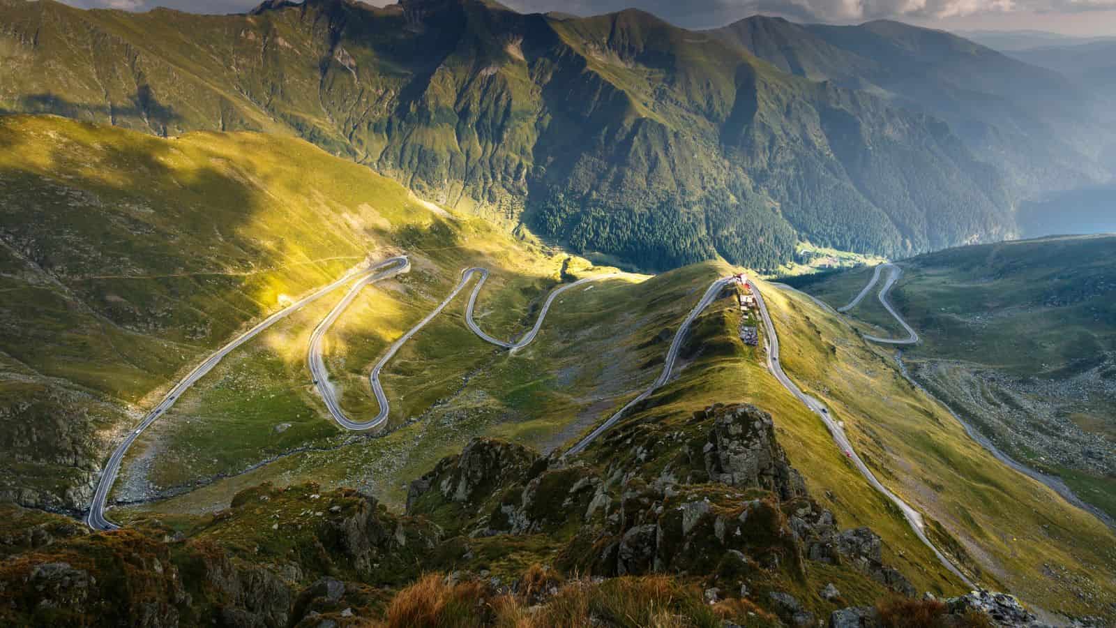 Winding mountain road through green hills under sunlight, with high peaks in the background.