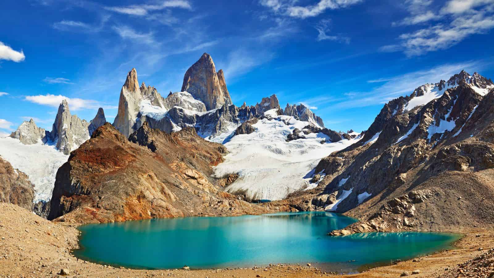 A turquoise lake with jagged, snow-capped mountains under a bright blue sky with scattered clouds.