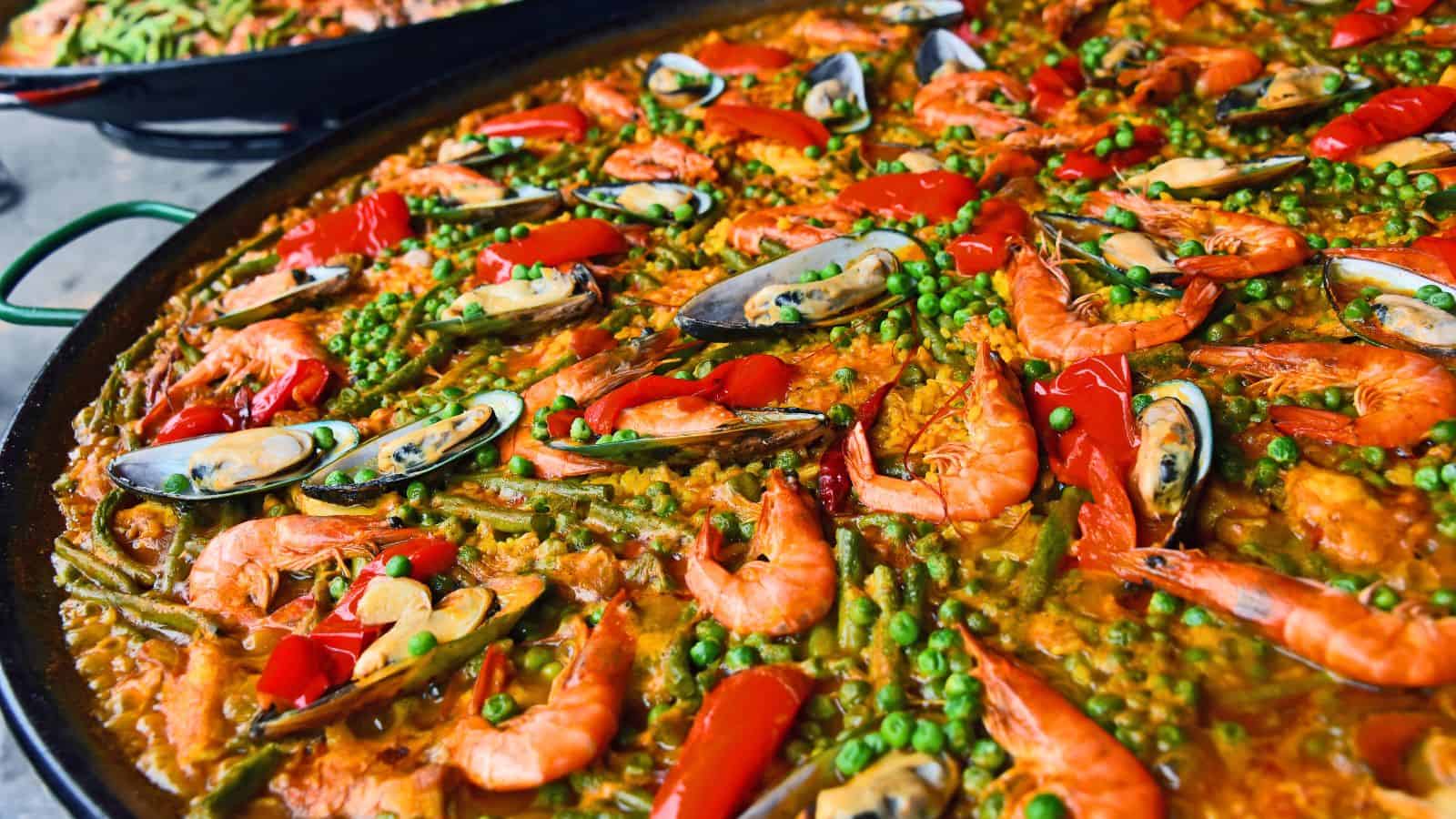 A large pan of paella topped with shrimp, mussels, green peas, green beans, and red bell pepper slices on a bed of yellow rice.