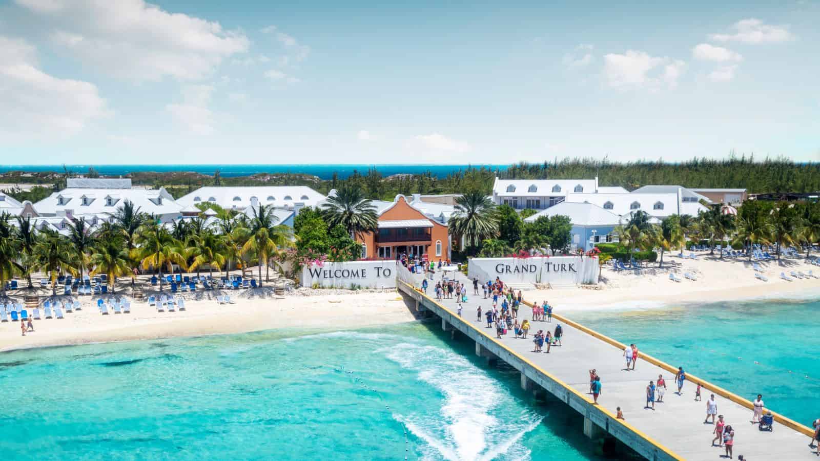A pier leads to a sunny beach with palm trees and a "Welcome to Grand Turk" sign, people walking ashore.