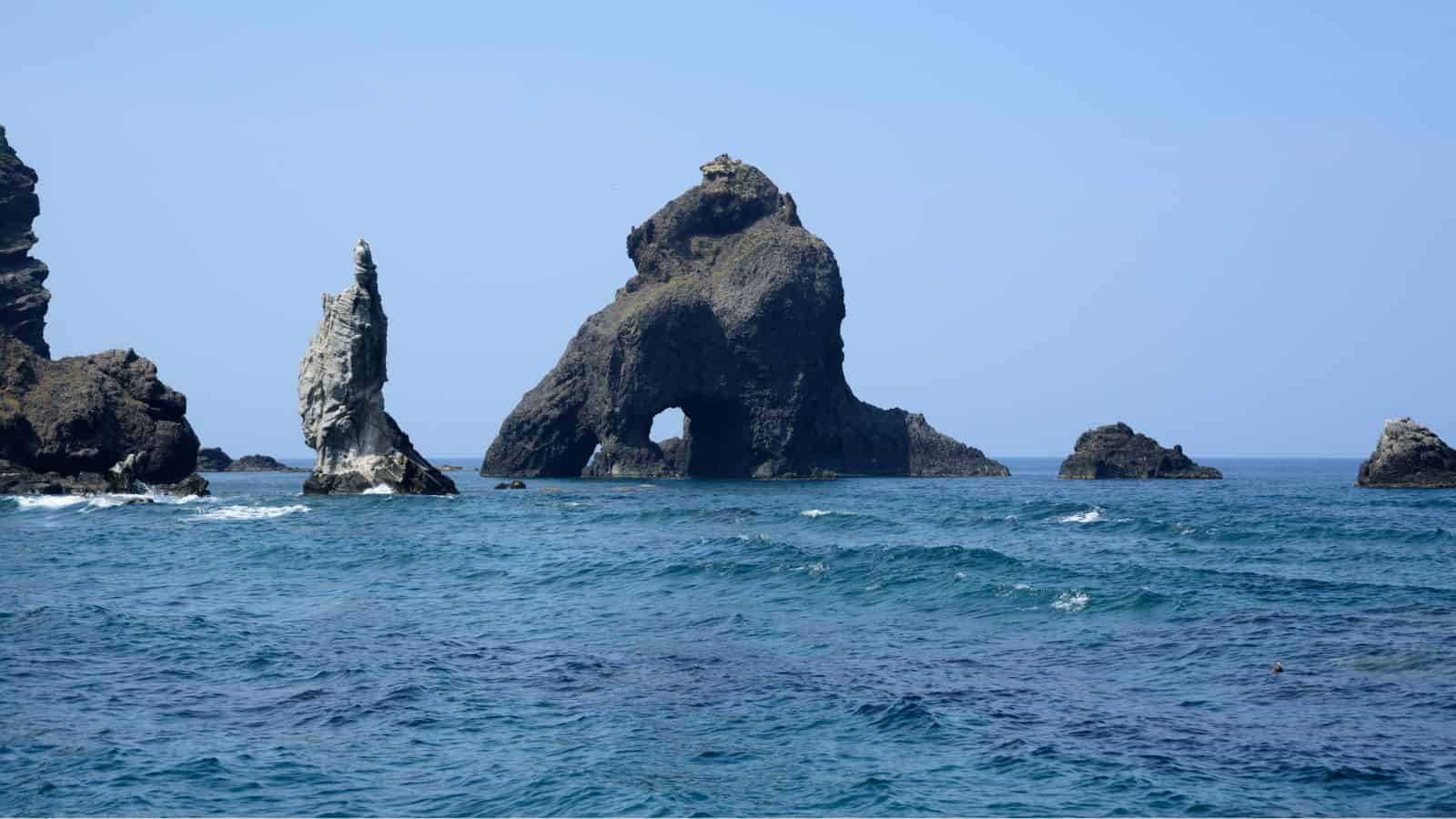 Rock formations rise from the blue ocean, with one arch-shaped rock in the center under a clear sky.
