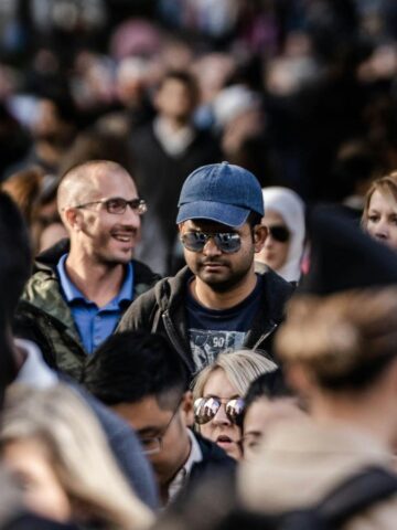 Crowded city street with many people walking, some wearing sunglasses, hats, and holding drinks.