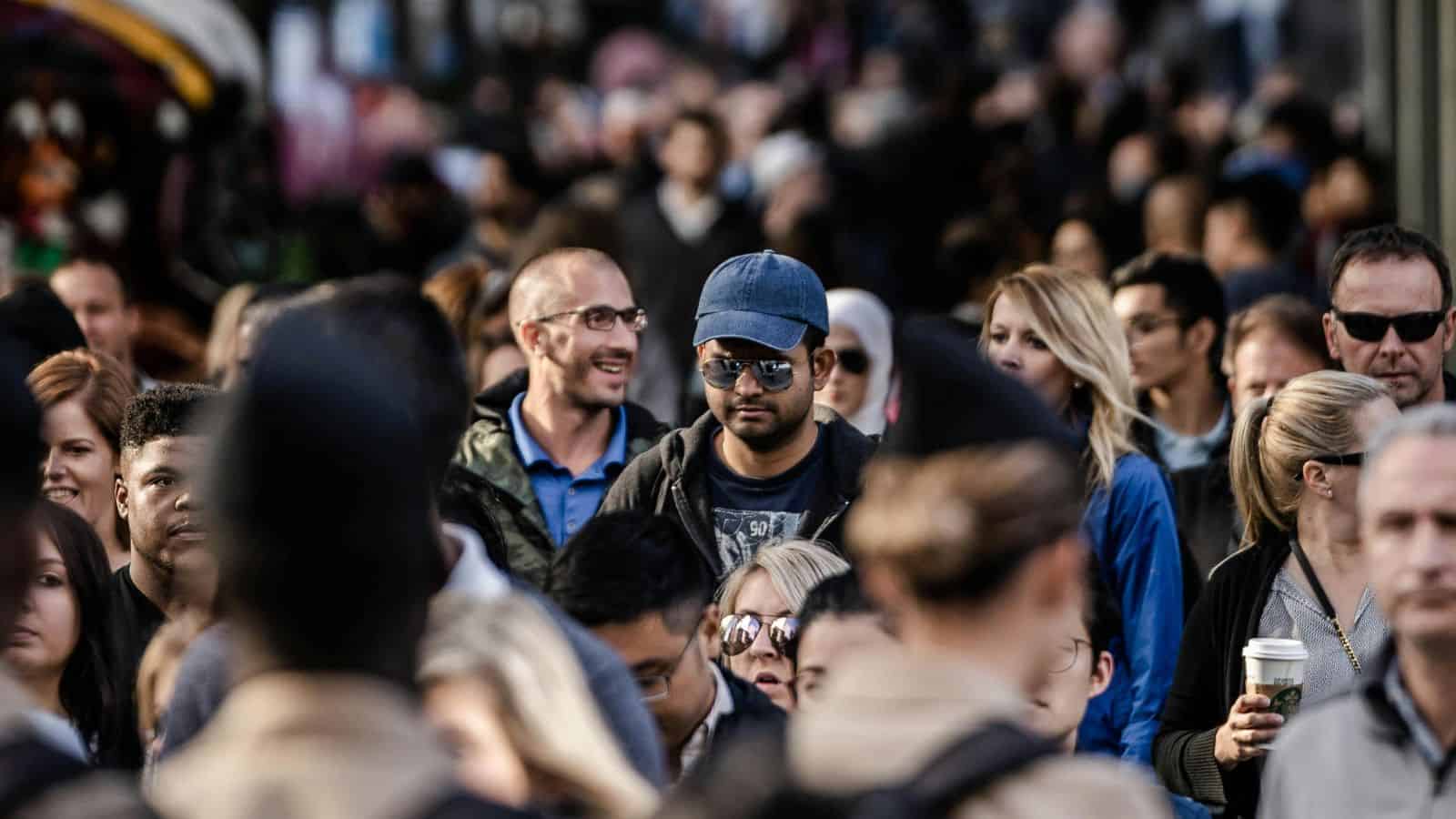 Crowded city street with many people walking, some wearing sunglasses, hats, and holding drinks.