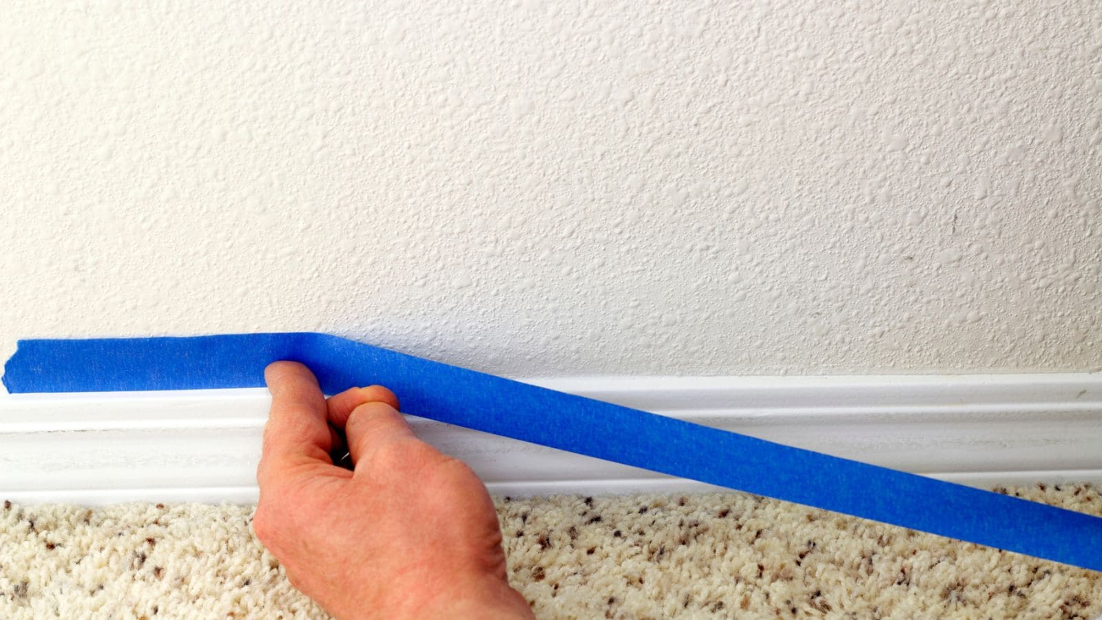 A hand applies blue painter&rsquo;s tape along a white baseboard above a beige carpet.