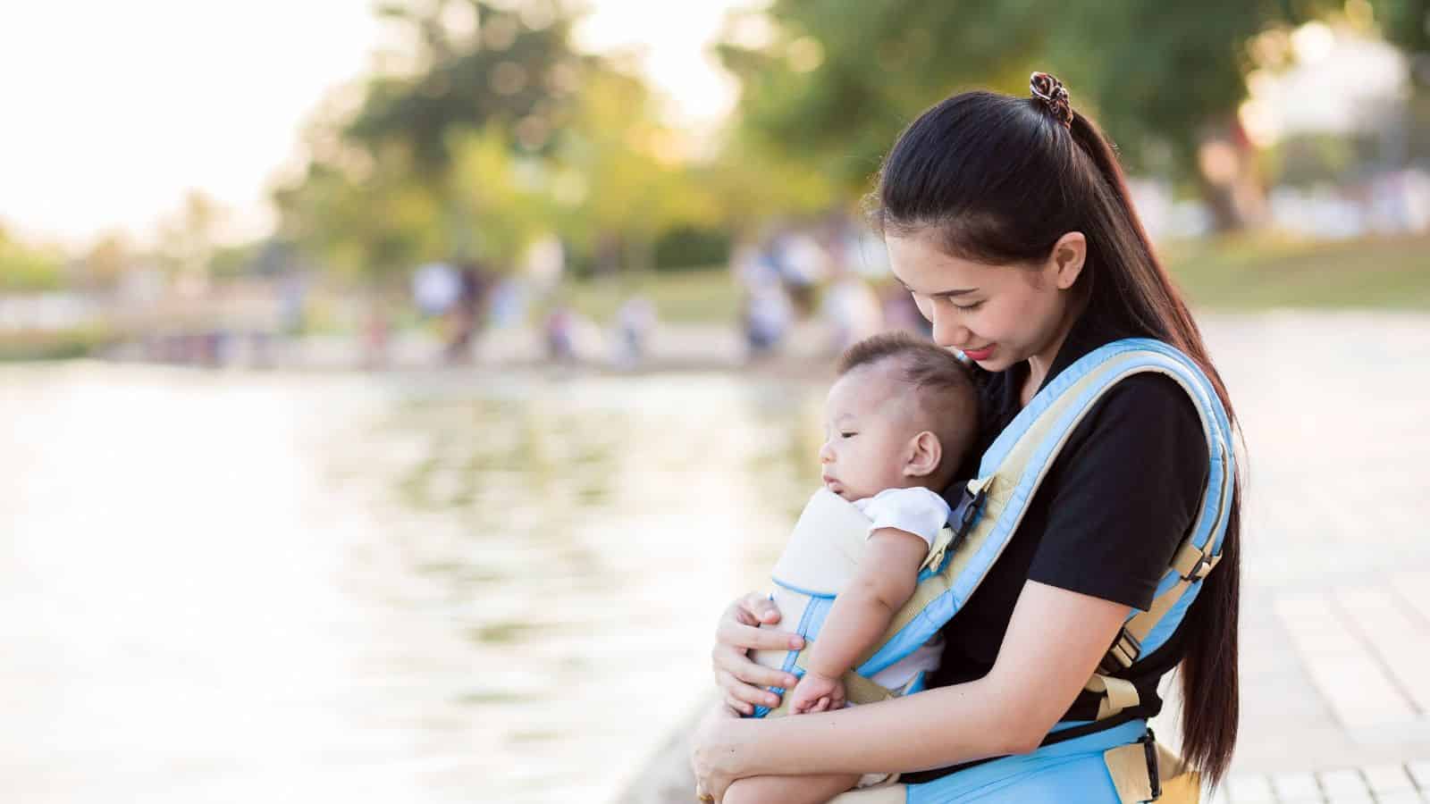 Woman smiling and holding a baby in a carrier by a lakeside path with trees in the background.