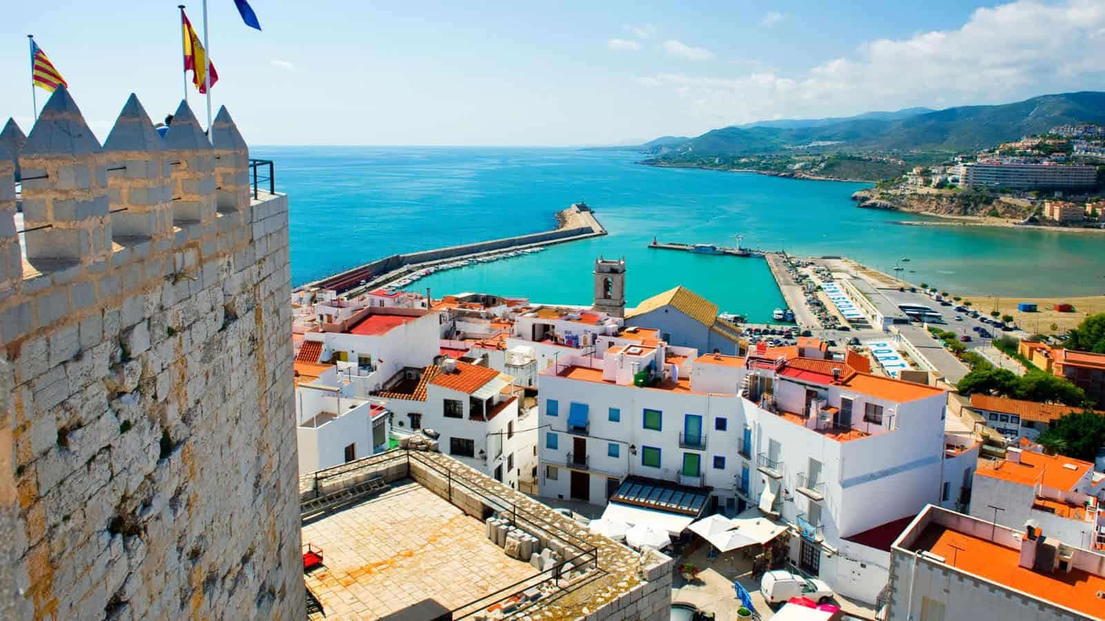 View of a coastal town with white buildings and red rooftops by a turquoise sea, a stone tower with flags, a marina, a pier, and hills in the distance under a blue sky with scattered clouds.