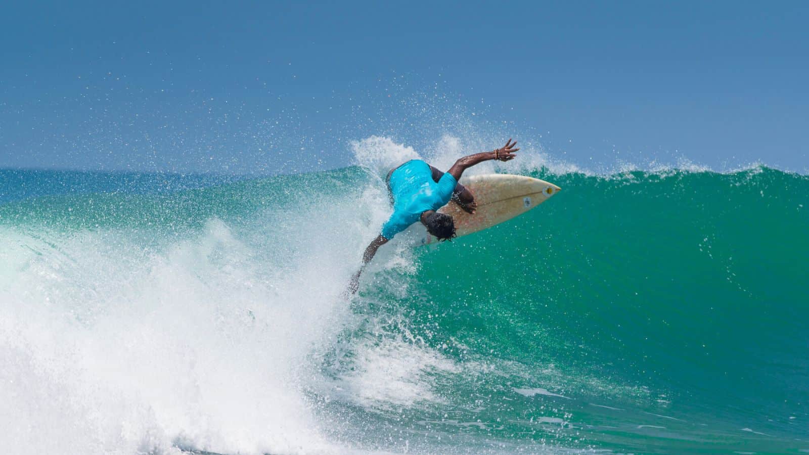 A surfer in a blue shirt rides a green wave at one of the top beach destinations with clear blue water, performing an aerial maneuver with one hand touching the sea and his surfboard angling above the crest under a clear sky.
