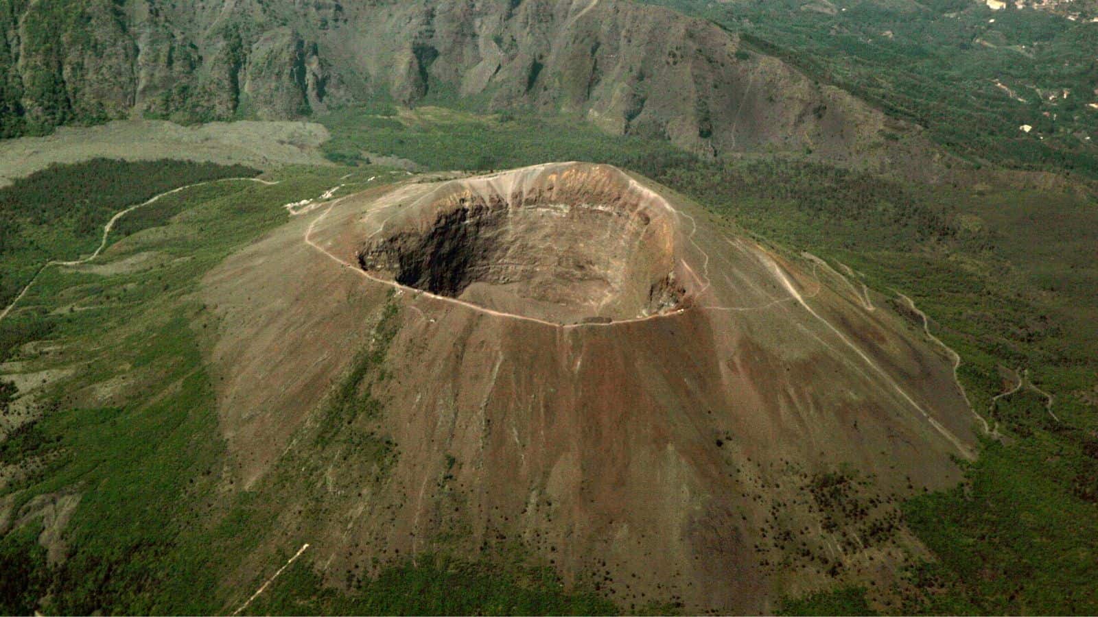 Aerial view of a large volcanic crater surrounded by rocky slopes and sparse vegetation, with mountains and greenery in the background.