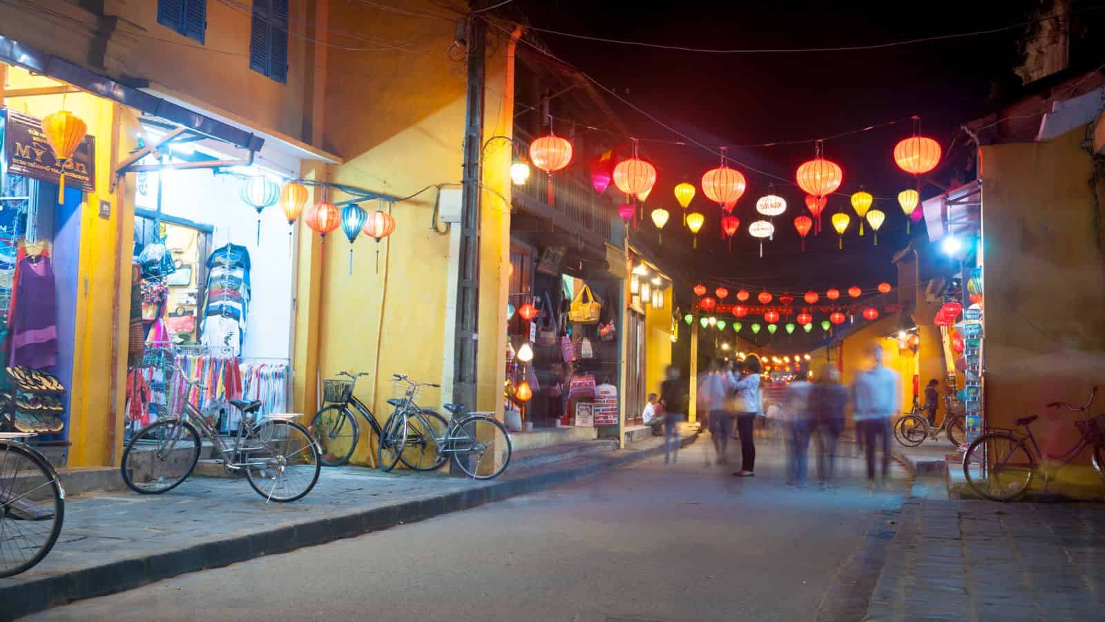 A vibrant street at night with colorful lanterns, bicycles, and blurred people walking by yellow buildings.