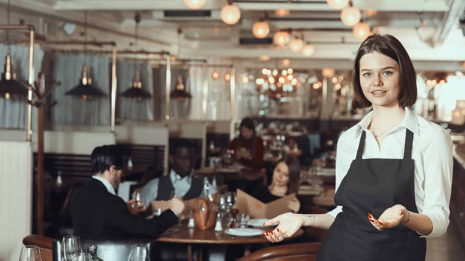 Smiling waitress in apron stands in restaurant with diners seated at tables in the background.