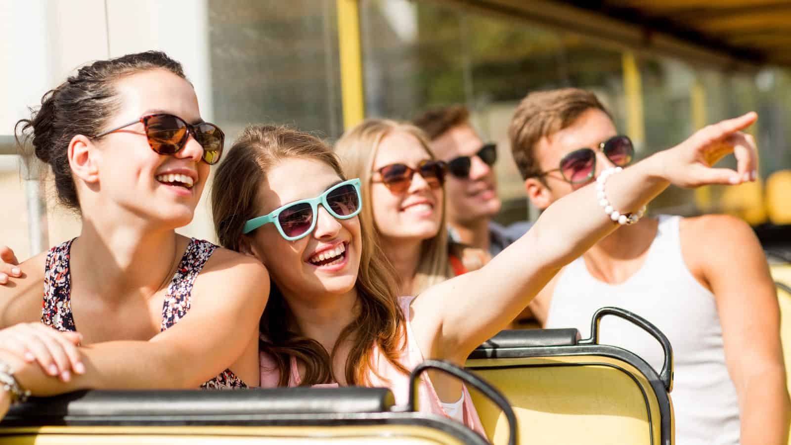 Five young adults wearing sunglasses smile and enjoy a ride on an open-top bus; one points excitedly ahead.