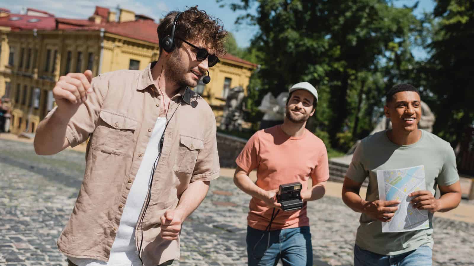 Three young men walk outdoors; one wears headphones, another holds a camera, and the third has a map.