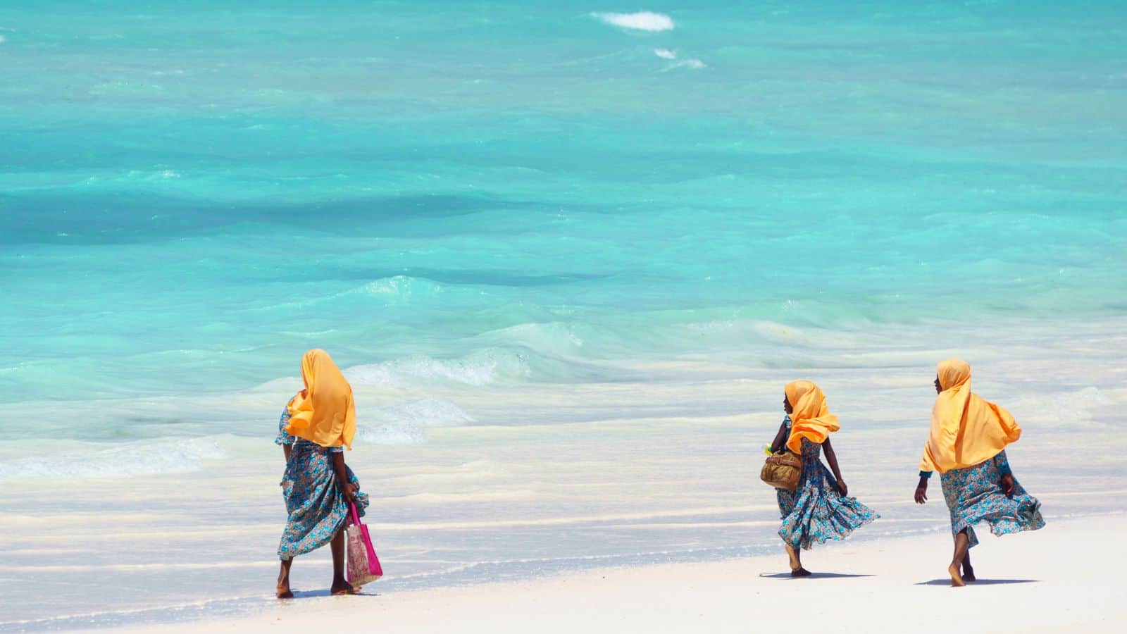 Three people wearing orange headscarves and patterned blue clothing walk along a white sandy beach beside turquoise ocean water under a bright sky.