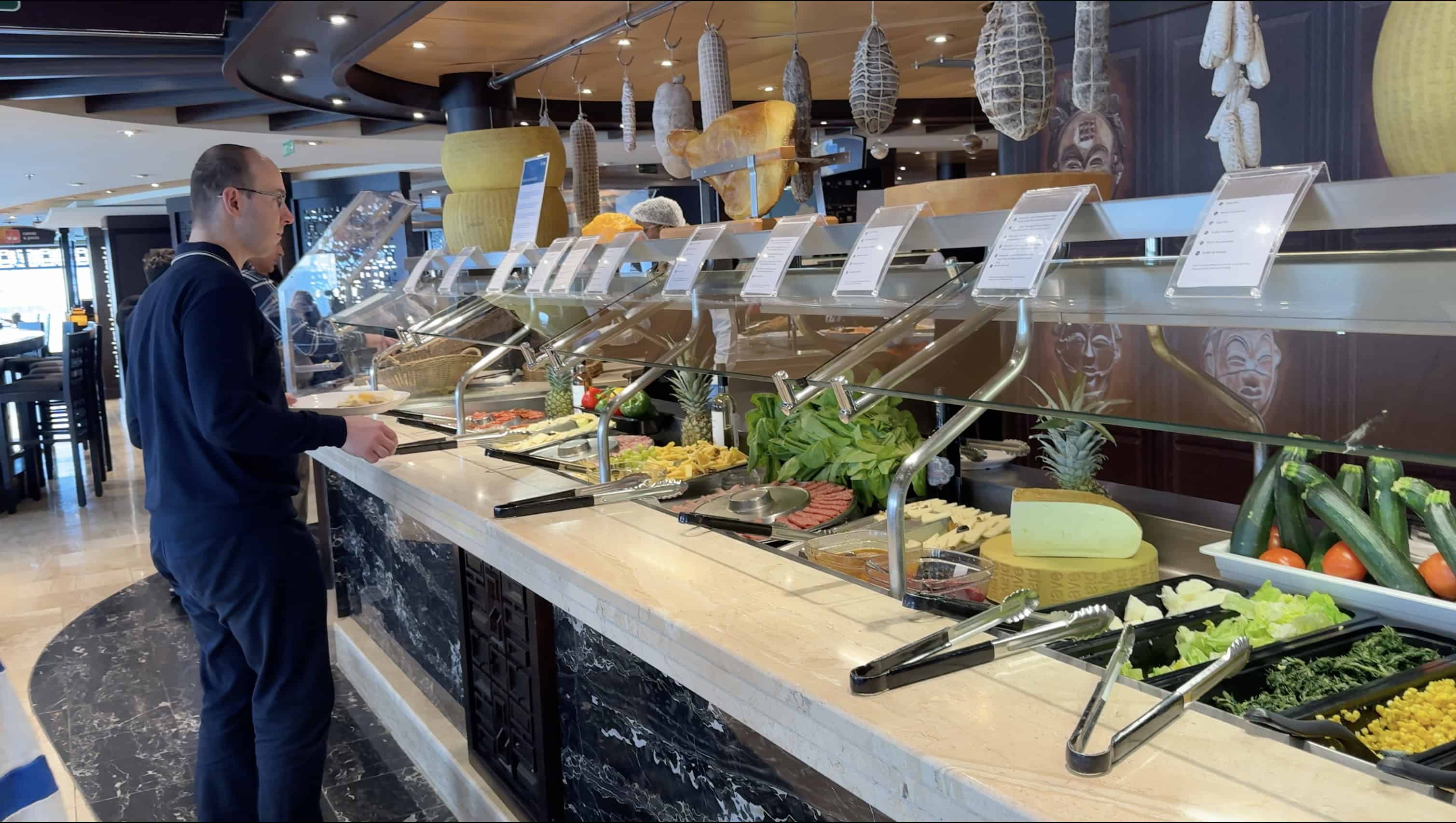 A man stands at a buffet on MSC Fantasia, with various salads, vegetables, and cheeses displayed under glass covers—perfect for a 2025 Cruise Review.