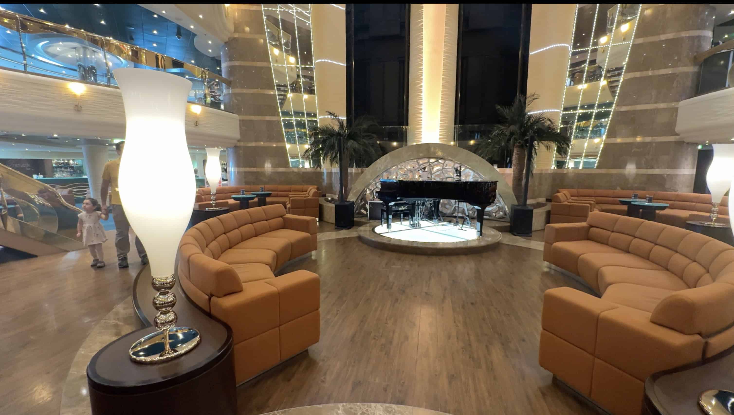 Luxurious lounge aboard MSC Fantasia with tan sofas, a grand piano on stage, tall lamps, and modern decor—perfect for relaxing evenings on your 2025 cruise. Read our Cruise Review for more highlights.