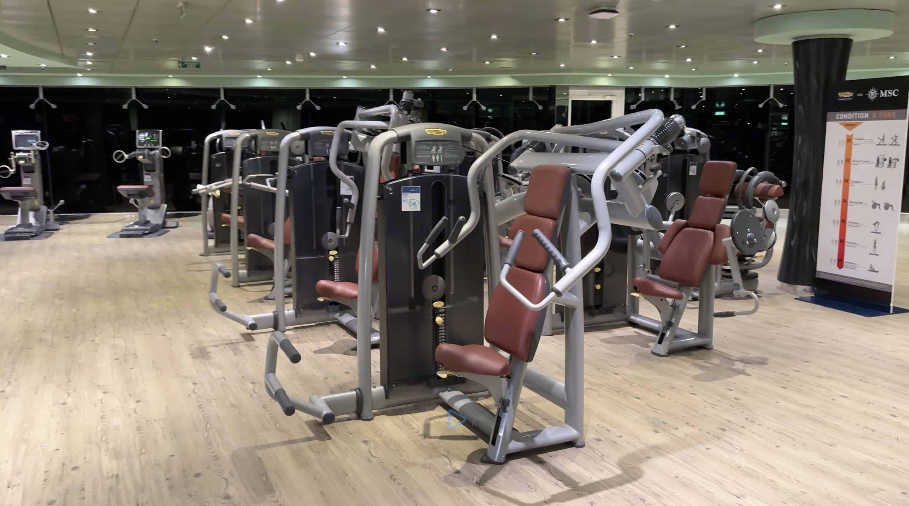 A modern gym with various weight machines and brown cushioned seats on a light wood floor, perfect for maintaining your fitness routine during your MSC Fantasia Cruise.