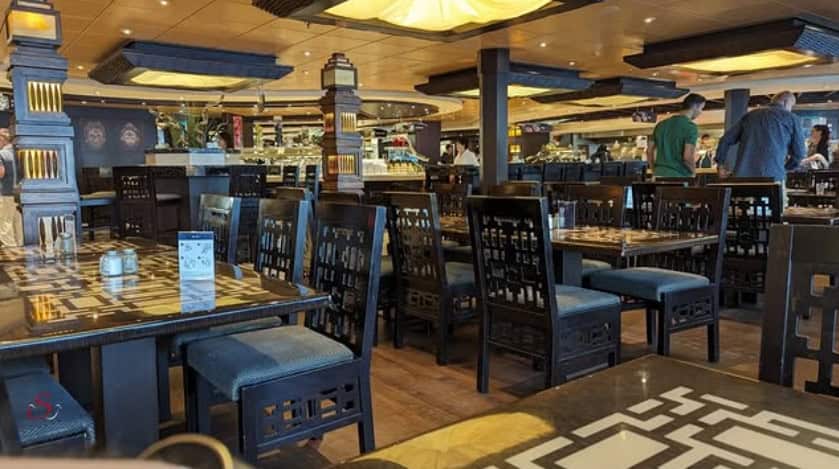 A modern restaurant interior inspired by MSC Fantasia Dining, featuring empty tables and chairs, with a few people standing near the buffet area.