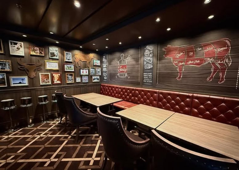 Cozy steakhouse with red leather seating, wood tables, cow art, and framed photos on dark paneled walls—evoking the refined charm of MSC Fantasia Dining.