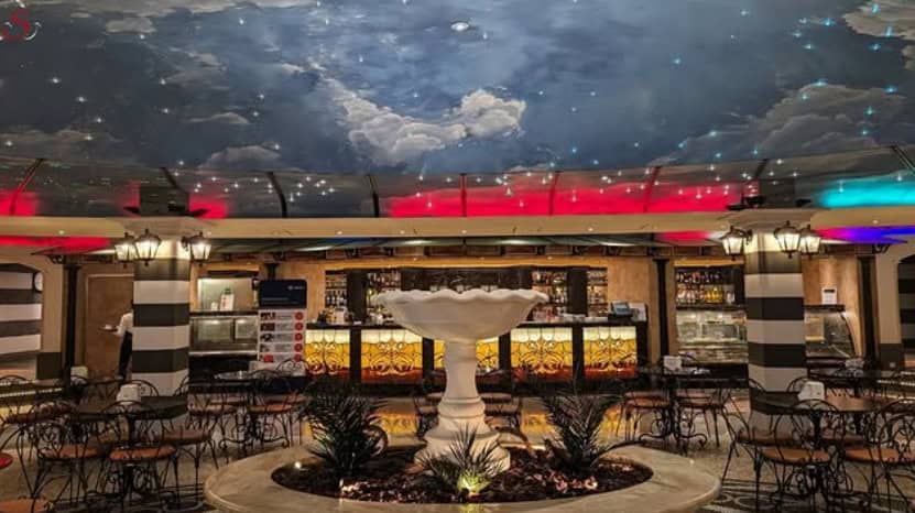 Indoor café with a central fountain, decorative ceiling lights, and tables surrounded by black chairs, reminiscent of the elegant MSC Fantasia Dining experience.
