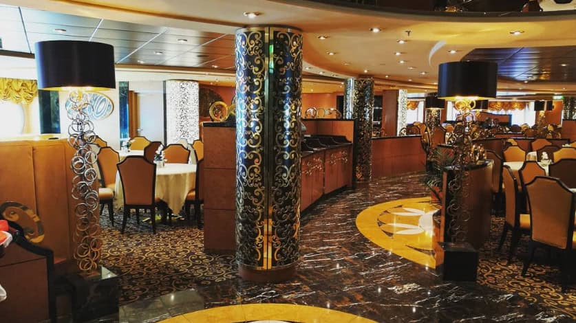 Elegant MSC Fantasia Dining room interior with ornate columns, gold accents, patterned carpet, and neatly arranged tables and chairs.