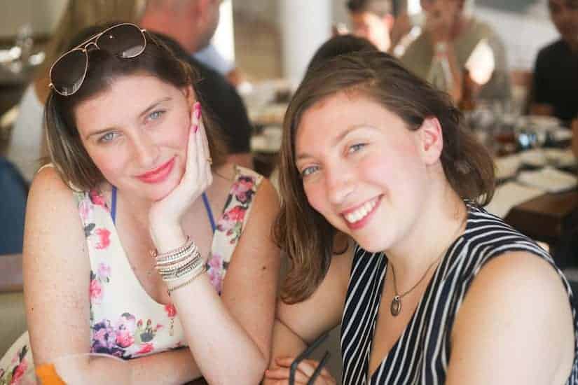 Two women smiling at a table in a bright, busy restaurant, one wearing a floral dress, the other in stripes.