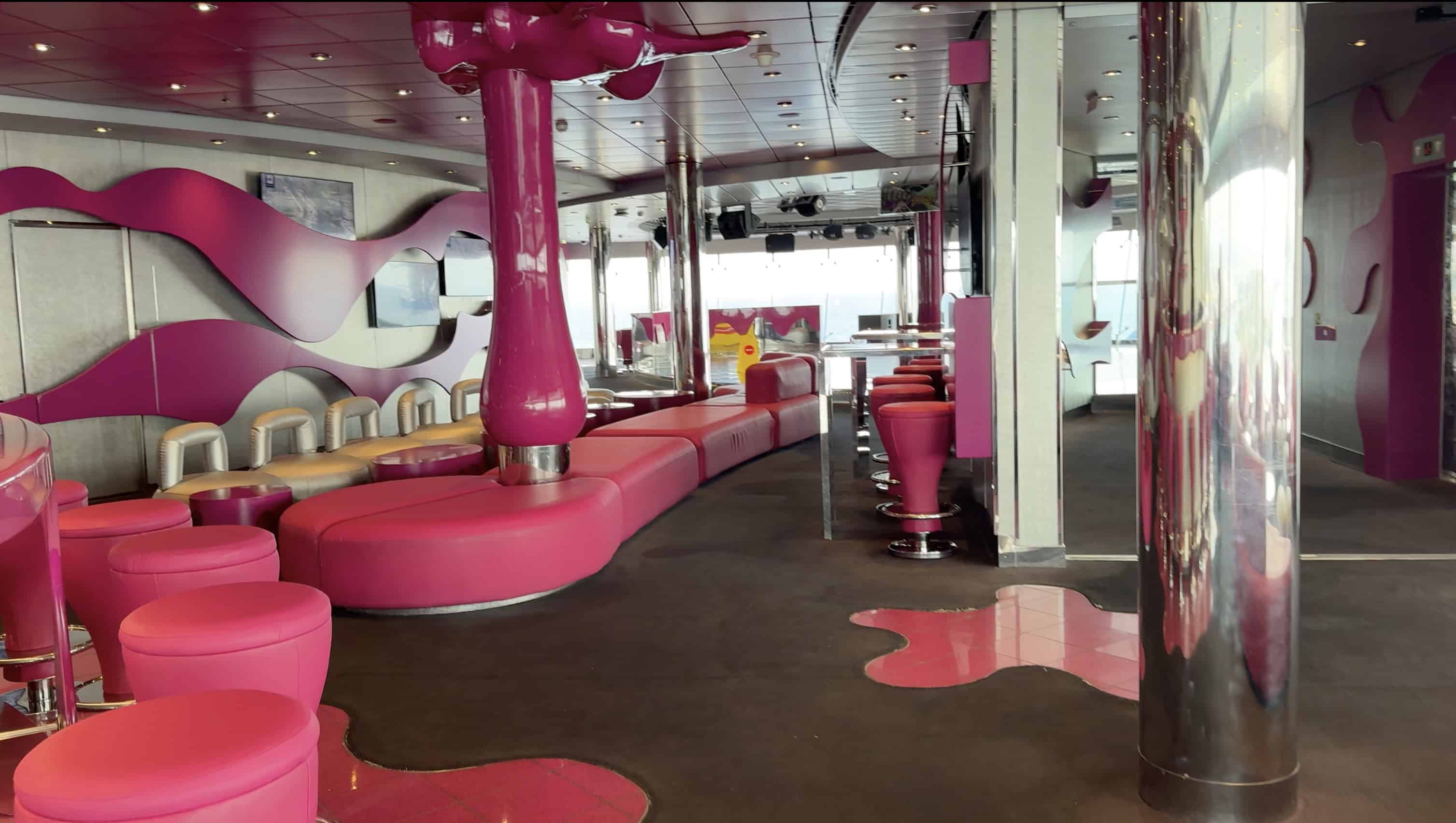 A modern lounge aboard MSC Fantasia with bright pink seating, curved sofas, bar stools, and wavy wall designs—perfect for your 2025 cruise review.