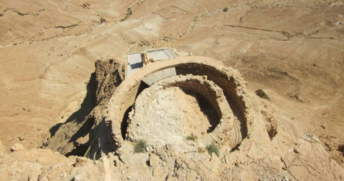 Ruins of a circular stone structure on a desert hillside with a small modern platform nearby, evoking the atmosphere of a trip to Masada.