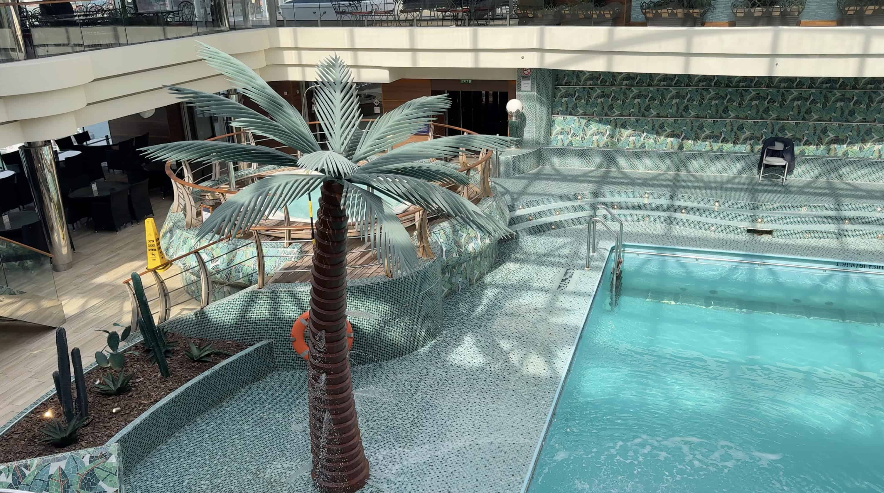Indoor pool area on the MSC Fantasia cruise features a fake palm tree, mosaic tile floor, and clear blue water—perfect for relaxing after a fun-filled family cruise.