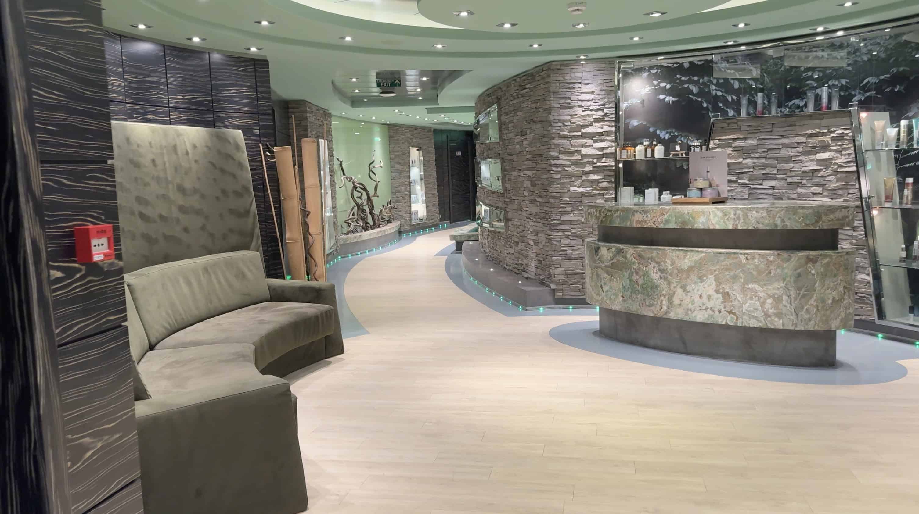 Modern spa lobby with stone walls, a curved green sofa, reception desk, and soft ambient lighting—perfect for relaxing before your MSC Fantasia Cruise. Start your Family Cruise Experience in style.
