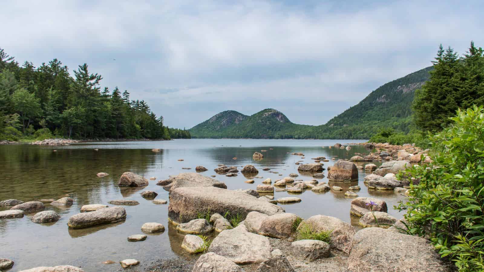 Rocky shoreline of a calm lake with forested hills and cloudy sky in the background.
