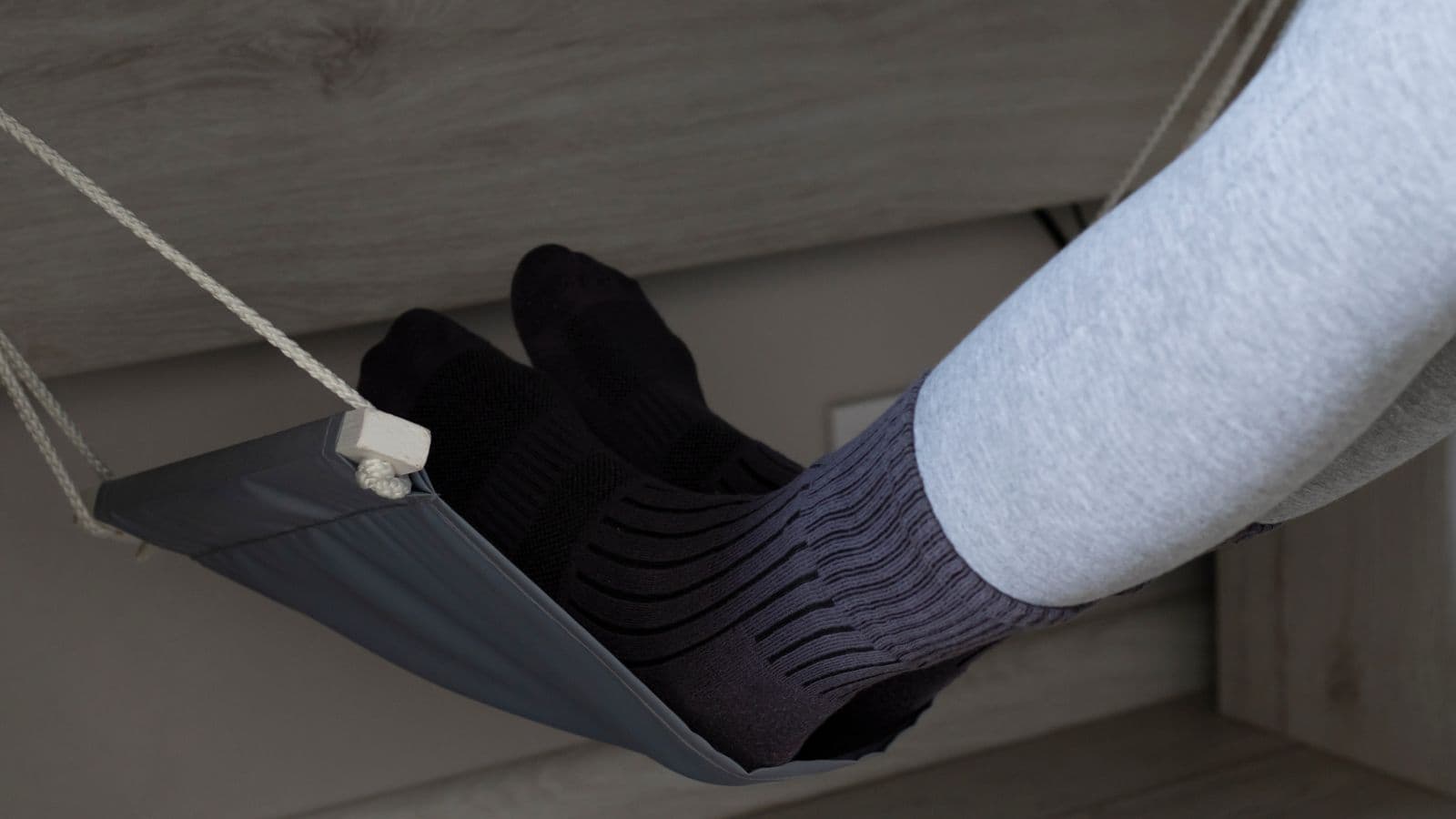 Person with gray pants and black socks resting feet on a small under-desk foot hammock, a handy travel product often used for sleeping on a plane.