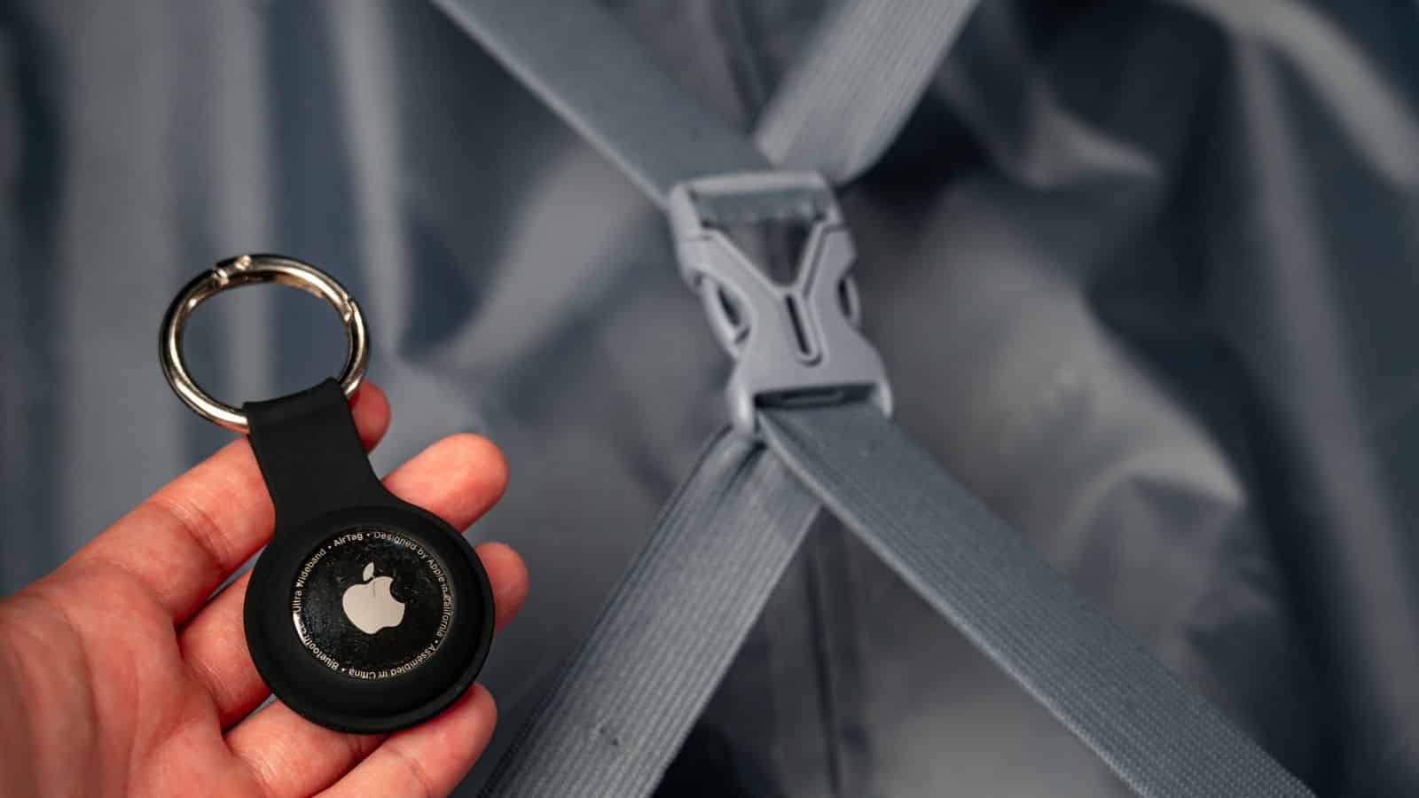 A hand holds an Apple AirTag with a keyring near blue suitcase straps and a plastic buckle.
