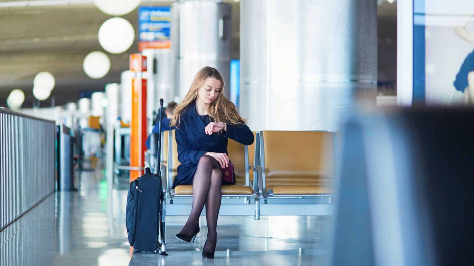 Woman in business attire sits in an airport, checking her watch with luggage beside her.